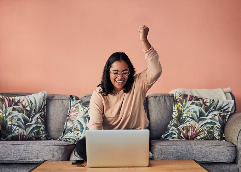 woman cheering while using a laptop on the sofa at home