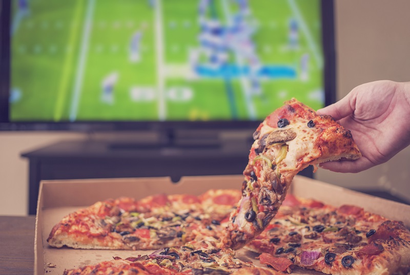 a person eating pizza out of a takeout box as they watch football