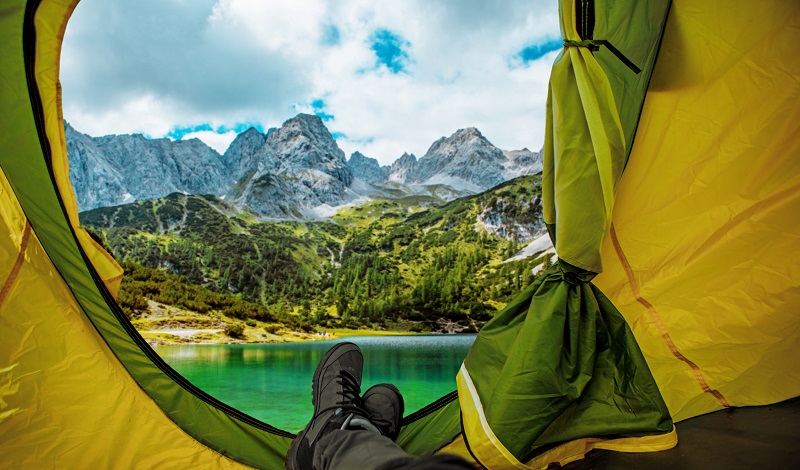 a person looking out of a tent, with a view of a lake, forest, and mountains