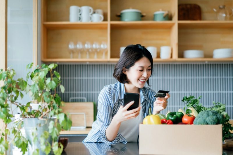 Woman ordering groceries online, with a box of fresh produce on the kitchen counter.