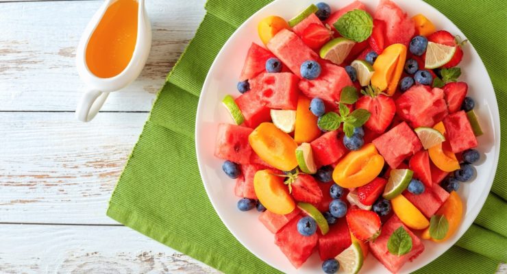 watermelon peach and berry salad with mint and lime wedges on a white plate on a wooden table with sweet orange dressing at the background, flat lay
