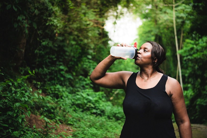 Woman walking alone through lush green forest path while drinking water