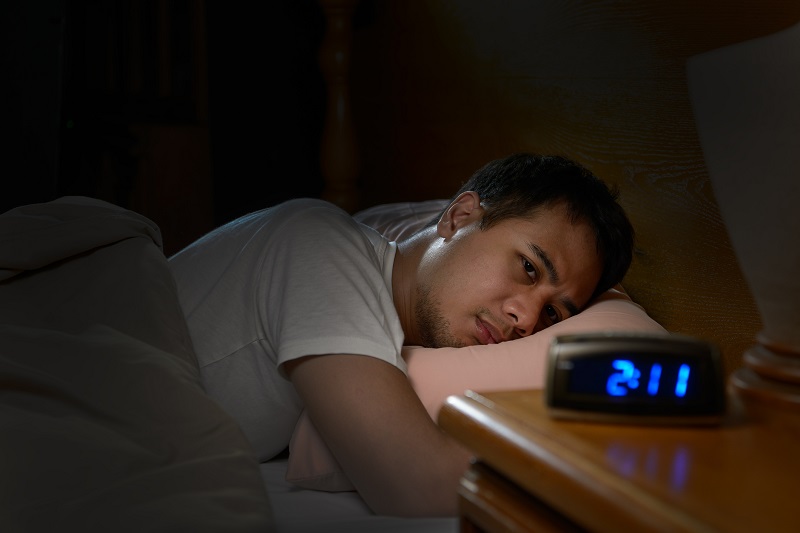 A man lies awake in bed and watches his alarm clock at night