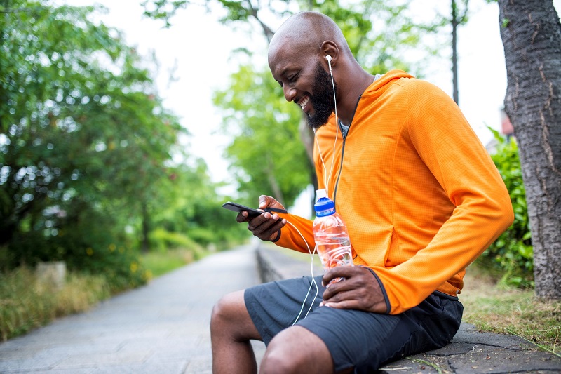 Man in athletic attire sitting on a ledge on a hiking trail, drinking water and checking his phone while smiling with earbuds