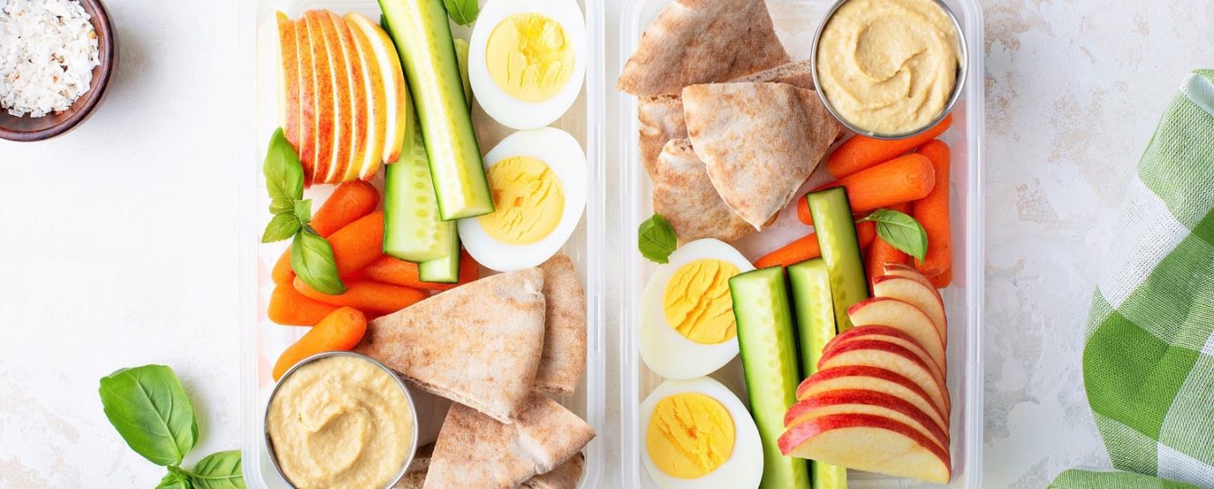 Healthy and nutritious snack boxes with veggies, pita bread, hummus, apple slices and a hard boiled egg