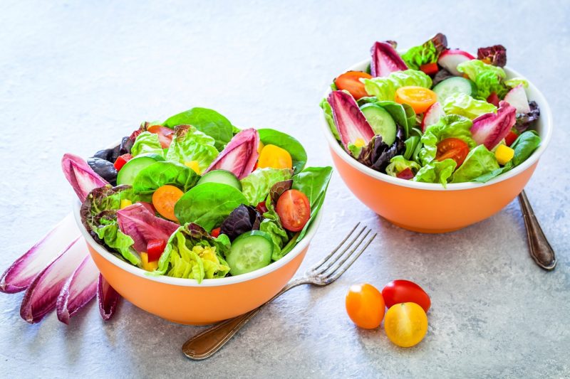 two healthy multi colored salad bowls on light gray table with lettuce, spinach, cherry tomato, arugula and cucumber.