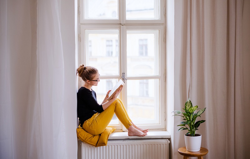 Woman perched on her windowsill, reading a book in the natural daylight