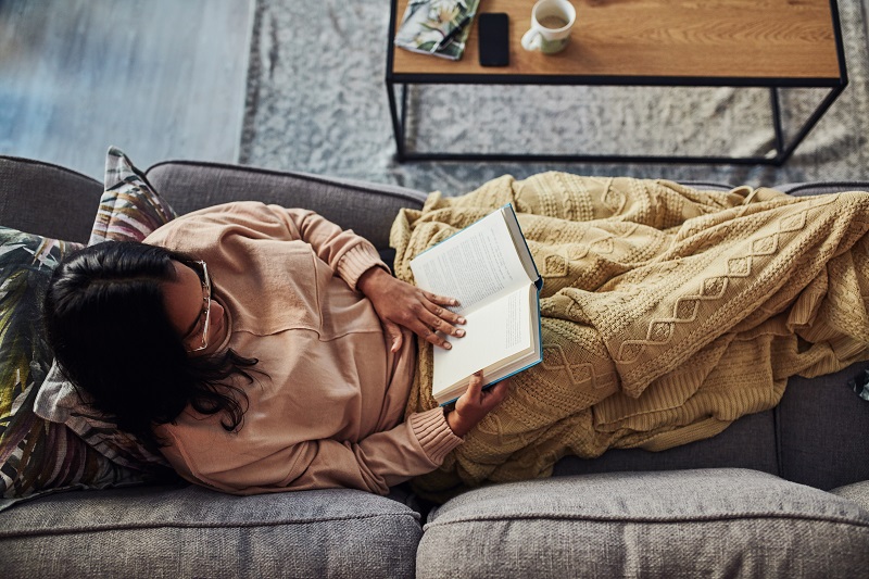 Woman reclines on couch, wrapped in a blanket, reading her book