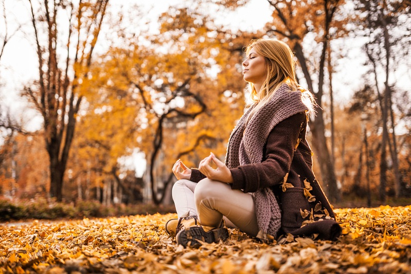 Self care for this woman meditates on the ground surrounded by yellow leaves and trees