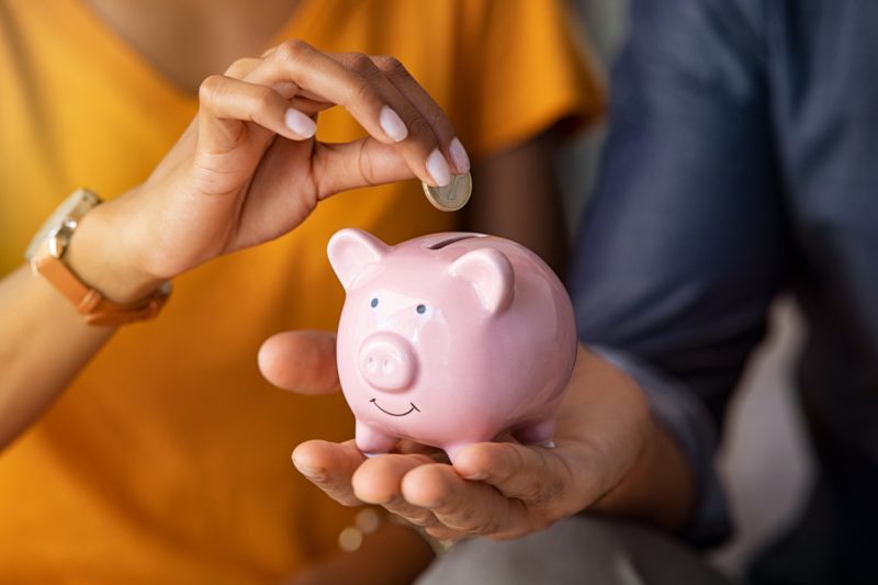 Close up of man holding pink piggy bank while woman putting coin in it.