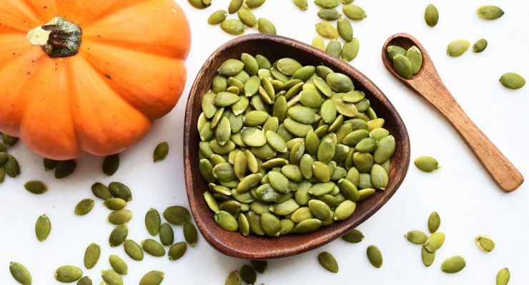 pumpkin seeds in a bowl next to a pumpkin and wooden spoon