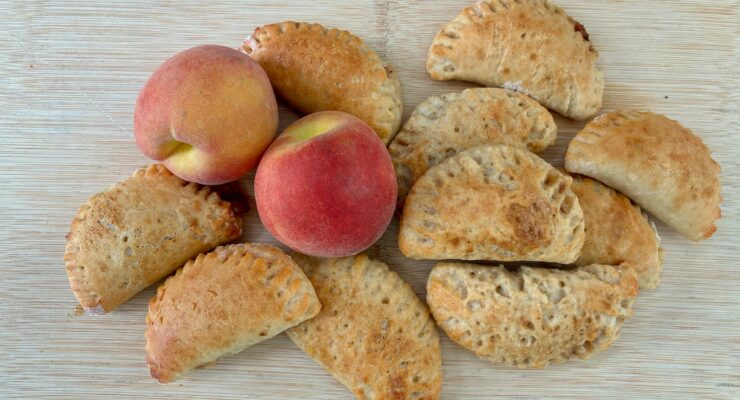 Golden-brown baked pastries and three ripe peaches on a wooden cutting board.